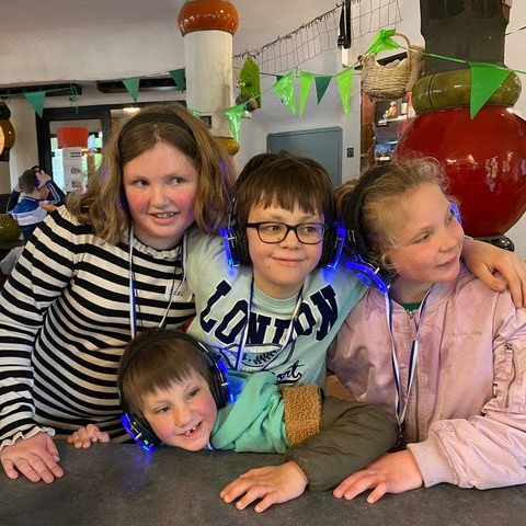 Vier kinderen met koptelefoons op, lachend, in een kamer versierd met vlaggen en grote fruitsculpturen.