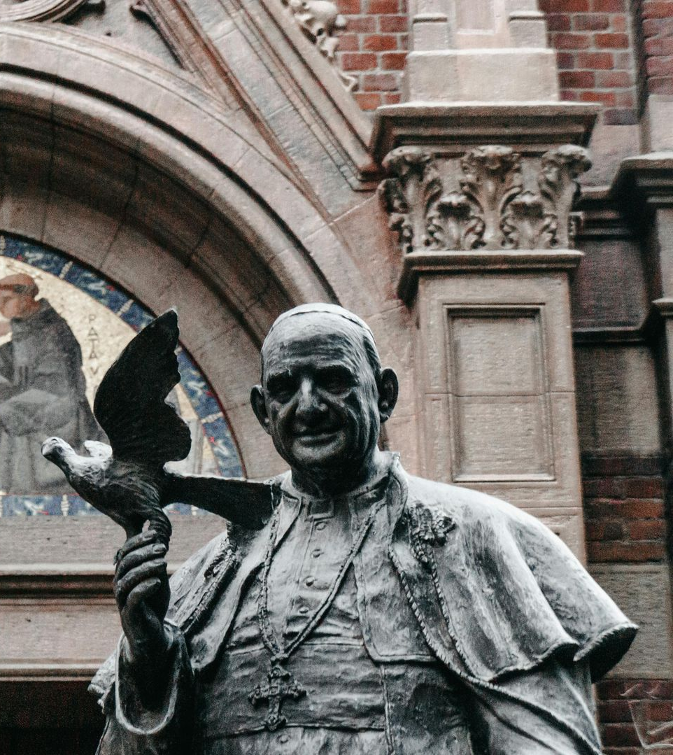 Statue of Pope John XXIII holding a dove in front of an arched building entrance.