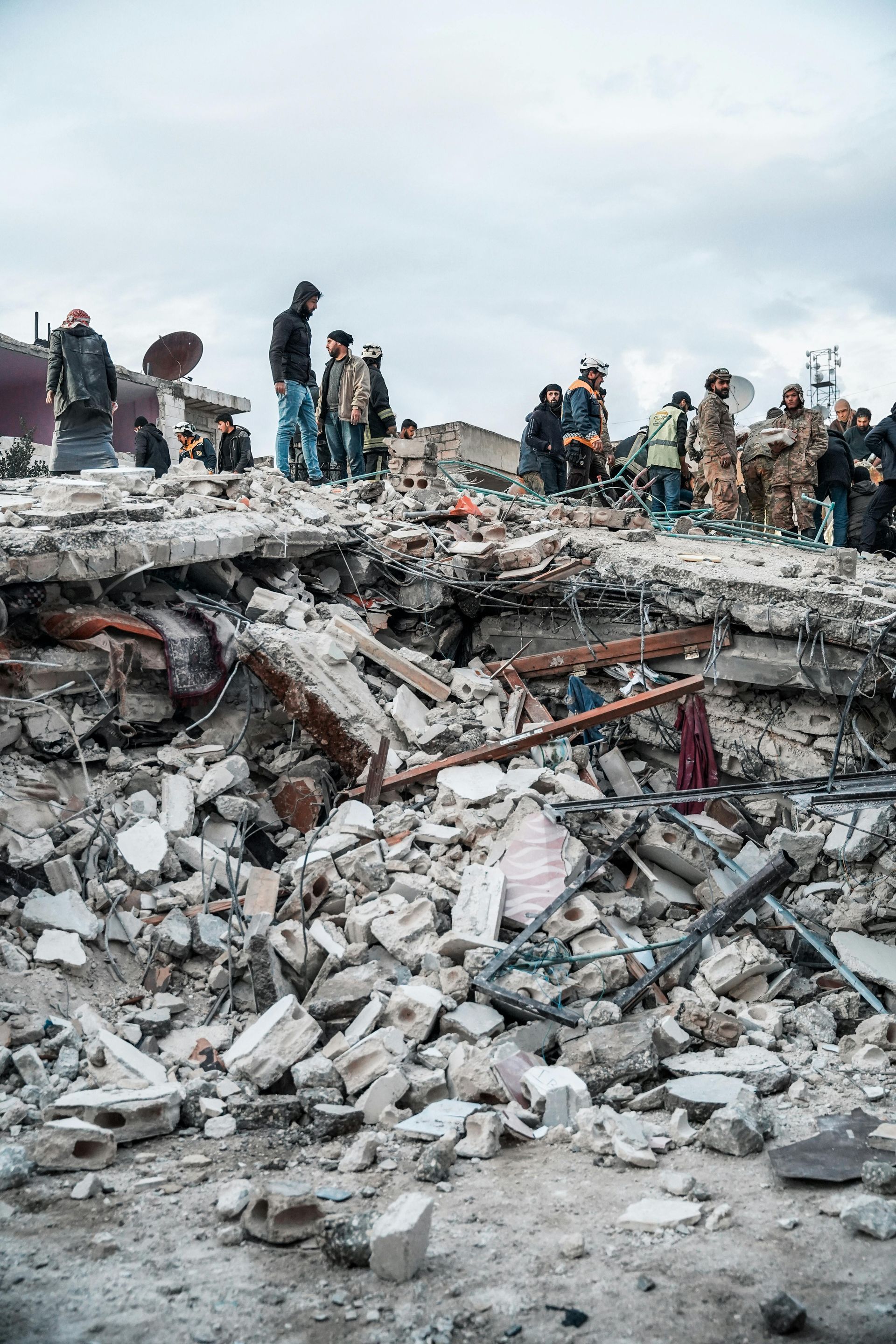 Debris and rubble from a collapsed building; people search for survivors amid the destruction under a cloudy sky.