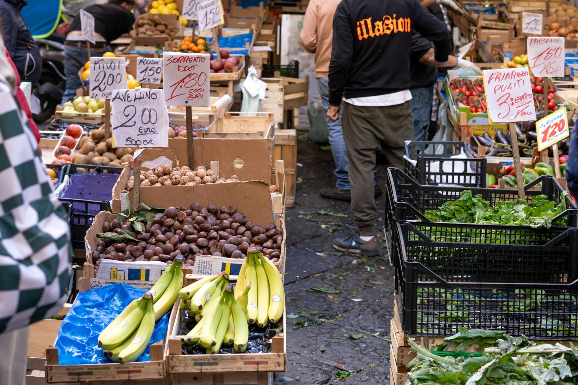 Produce stand with produce and customers. Bananas, greens, and other fruits are on display.