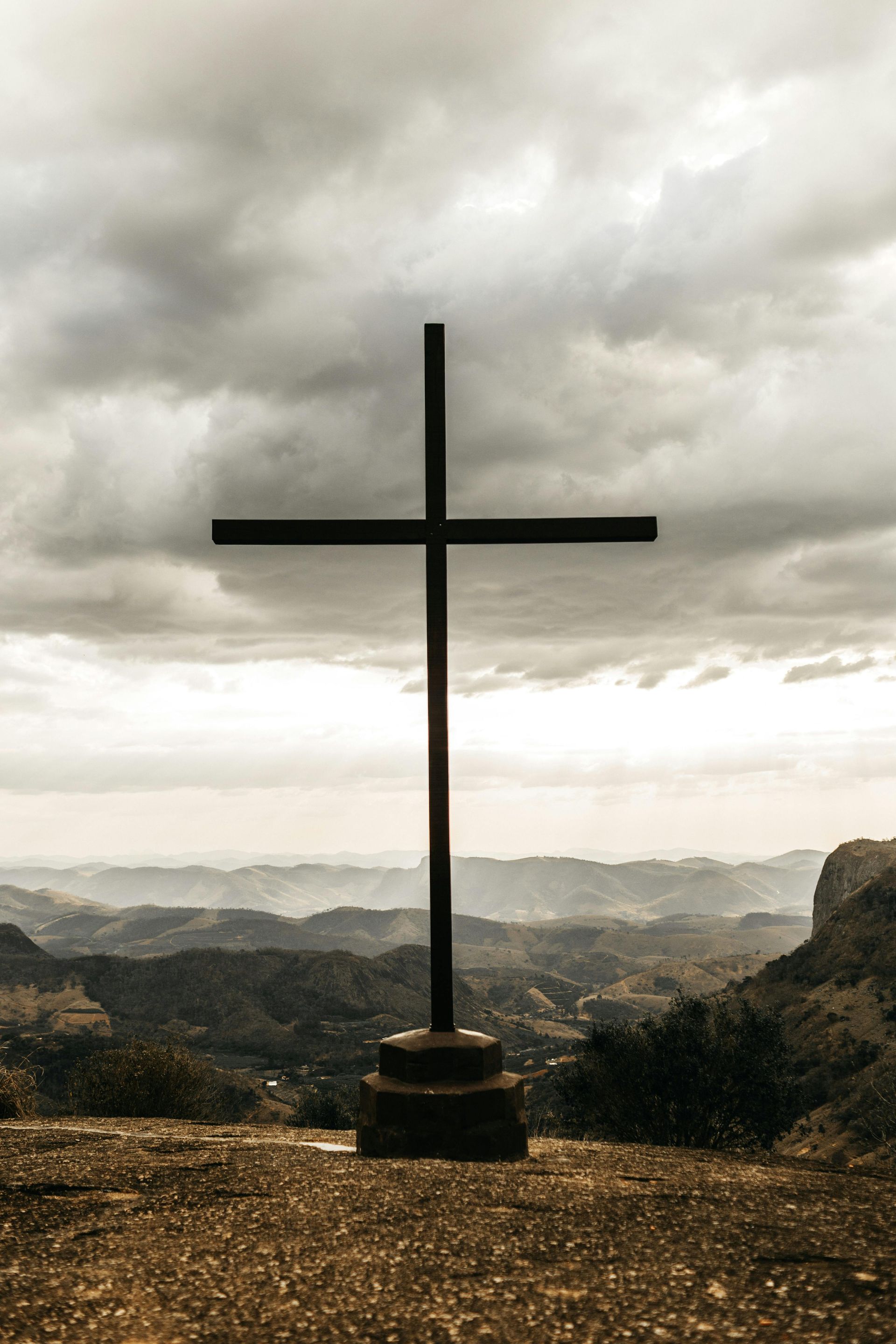 Cross on a hilltop silhouetted against a cloudy sky, overlooking distant mountains.