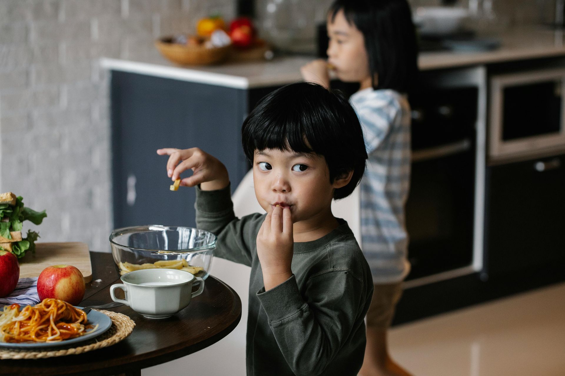Boy in green shirt eating snacks from bowl in kitchen, girl in background.