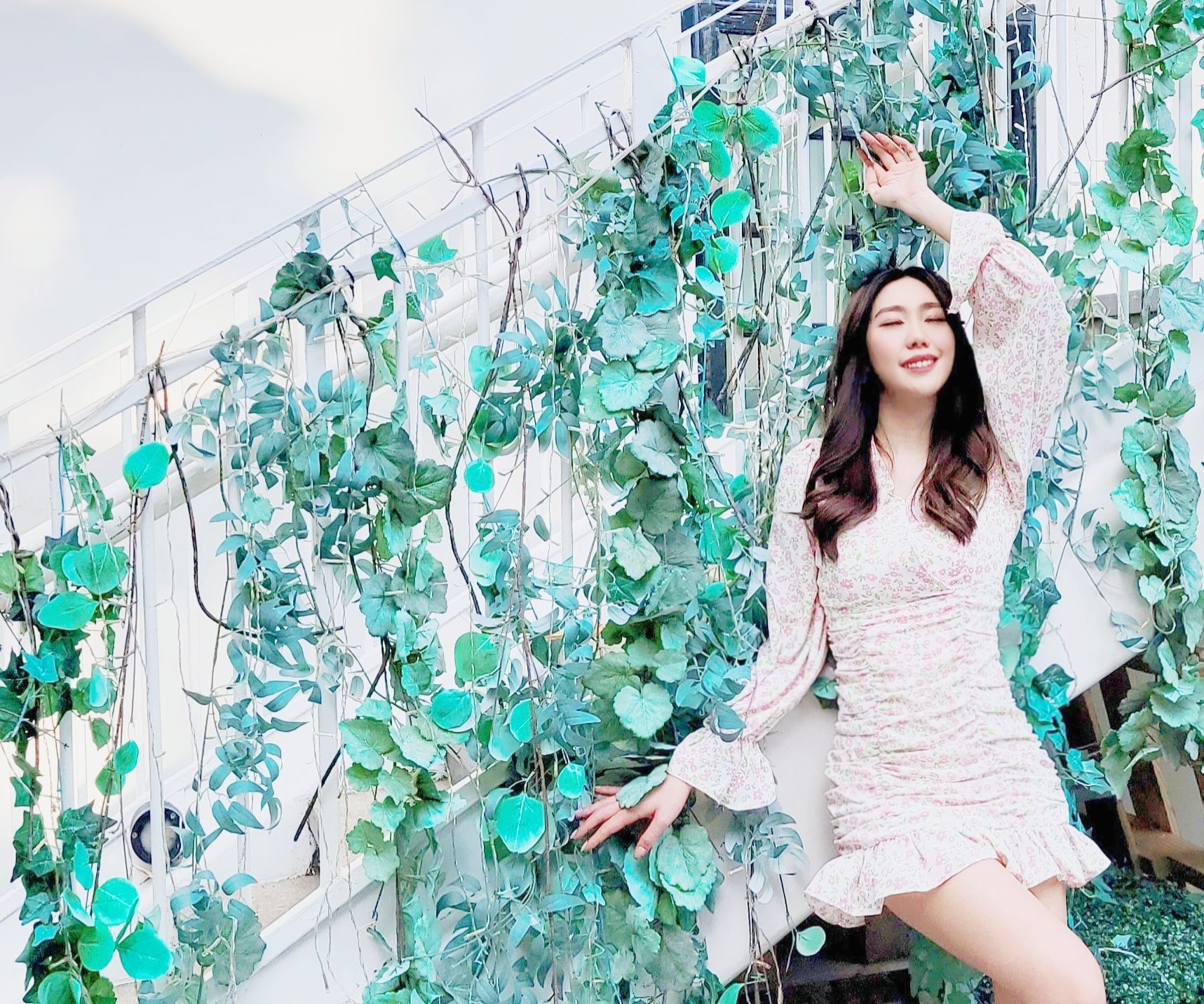 A woman in a white dress is standing in front of a wall of green leaves.