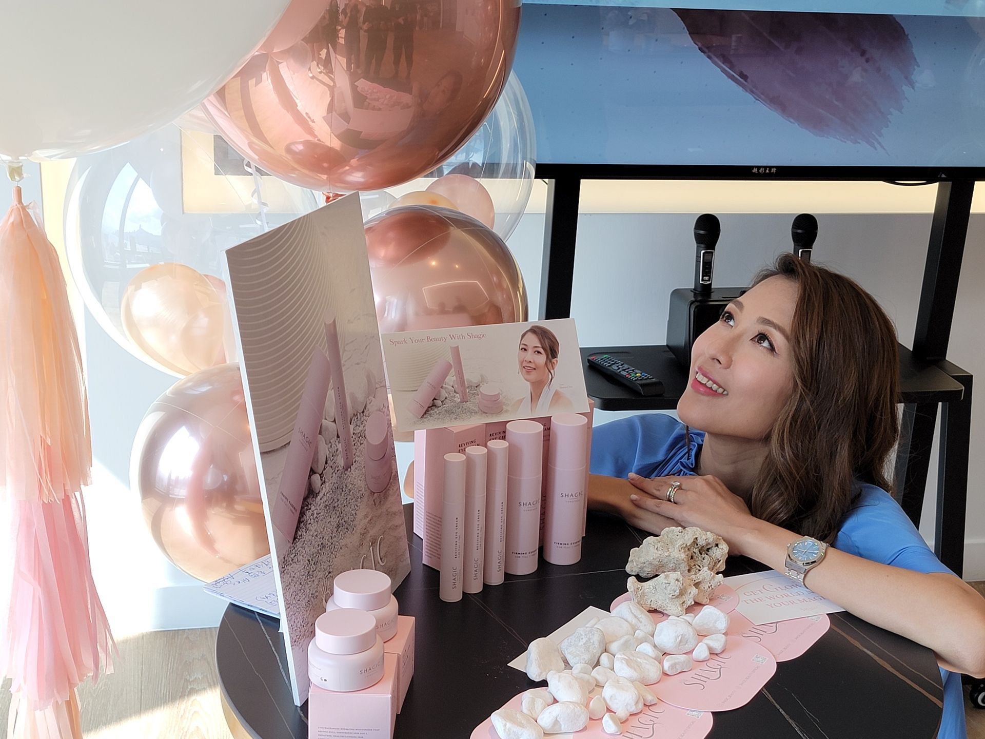 A woman is sitting at a table with balloons and cosmetics on it.