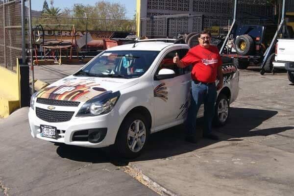 Un hombre está de pie junto a un sedán Chevrolet blanco con calcomanías de carreras en un estacionamiento.