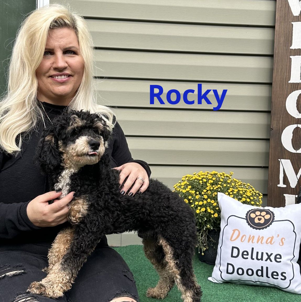 A woman is sitting next to a dog and a pillow with the name rocky on it.