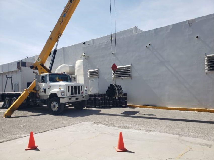 Una grúa eleva objetos cerca de un edificio blanco con rejillas de ventilación. Conos de tráfico naranjas en primer plano.