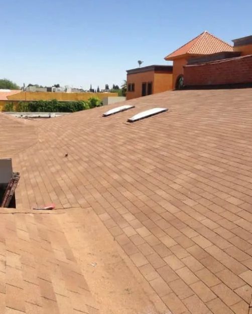 Techo de tejas asfálticas marrones con dos tragaluces, edificio al fondo bajo un cielo azul.