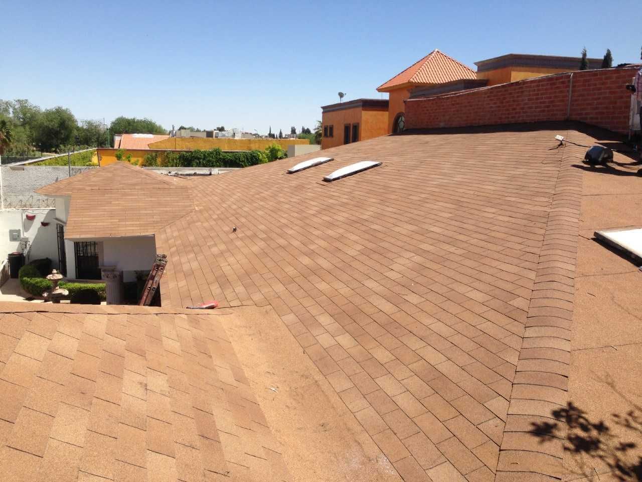 Tejado de tejas marrones con tragaluces en un día soleado. Edificios al fondo con paredes de terracota y blancas.