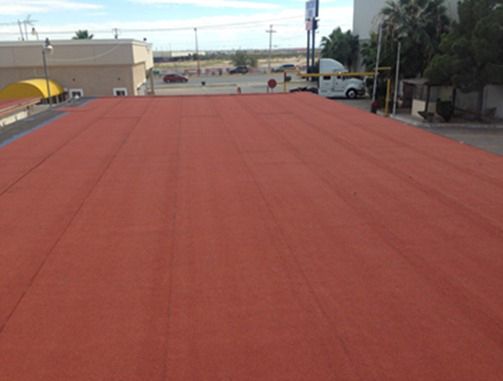 Techo plano rojo de un edificio, con algunos otros edificios y coches al fondo en un día soleado.