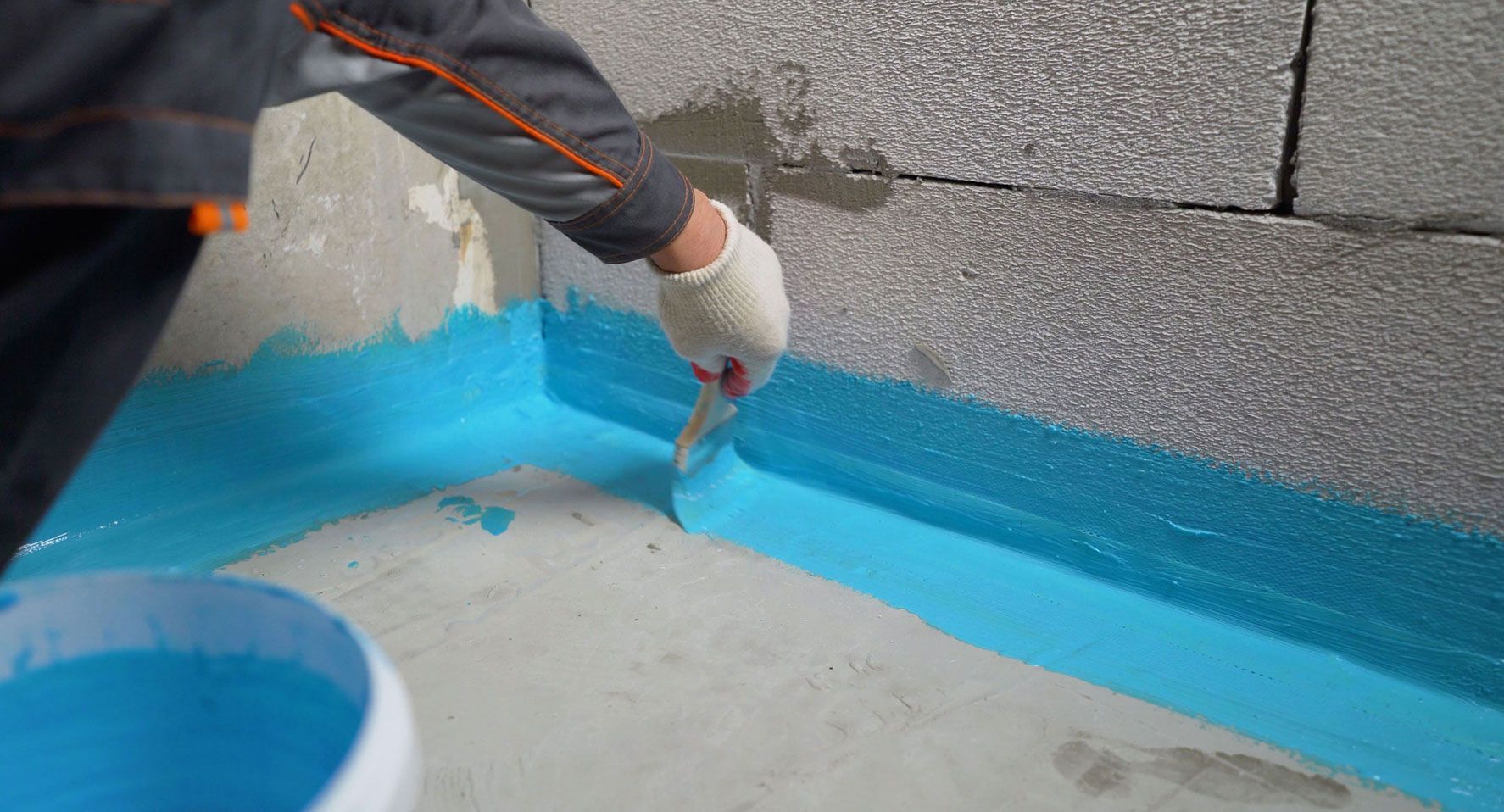 Una persona aplica sellador azul en la esquina de una pared. La escena transcurre en una obra en construcción, con un cubo de sellador a la vista.