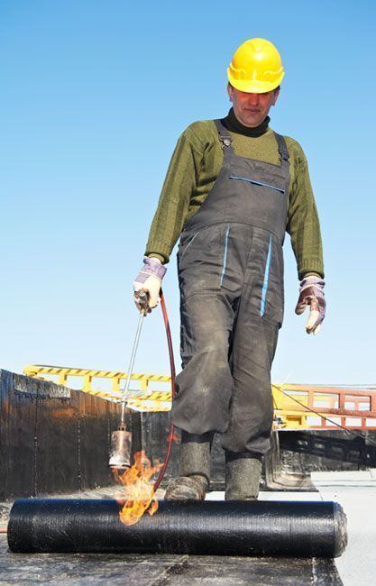 Un techador con casco amarillo utiliza un soplete para calentar un rollo de material para techos en una azotea plana.