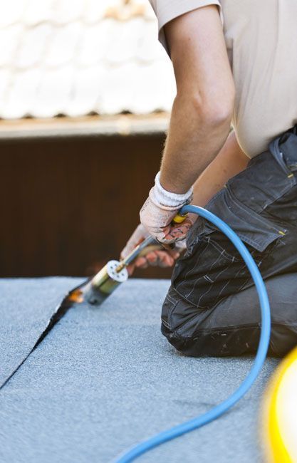 Techador arrodillado, utilizando un soplete para instalar material de techado; manguera azul conectada.