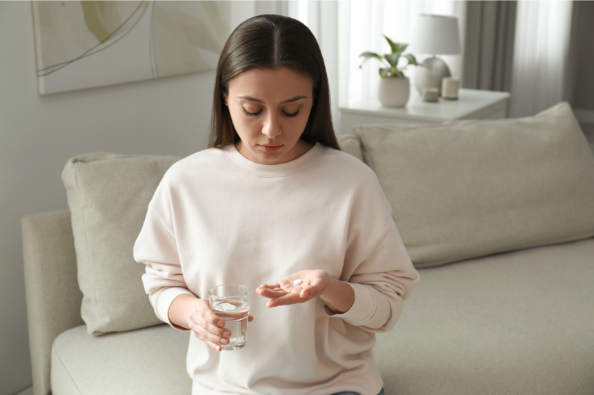 A woman is sitting on a couch holding a glass of water and a pill.