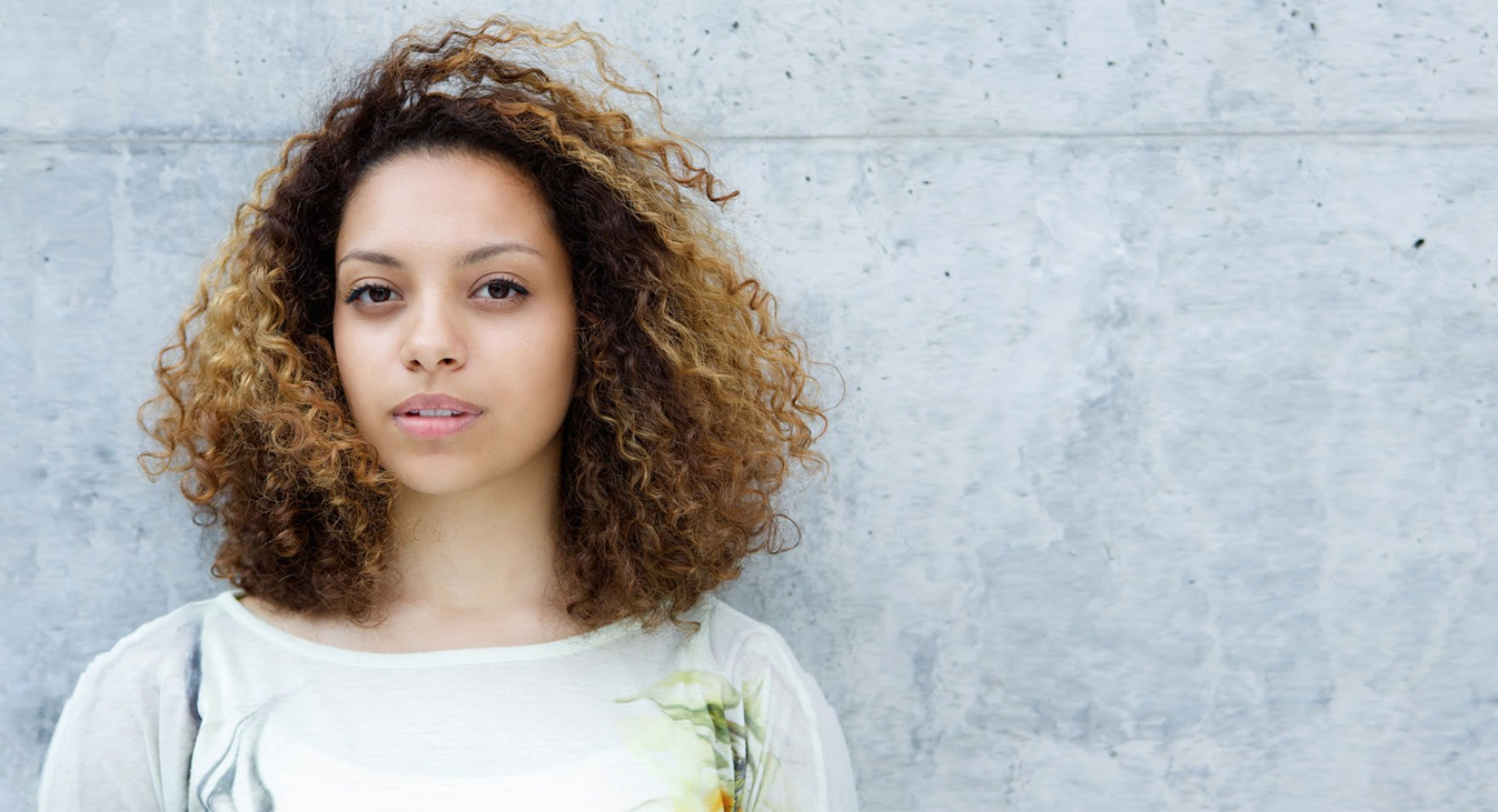 A woman with curly hair is standing in front of a concrete wall.