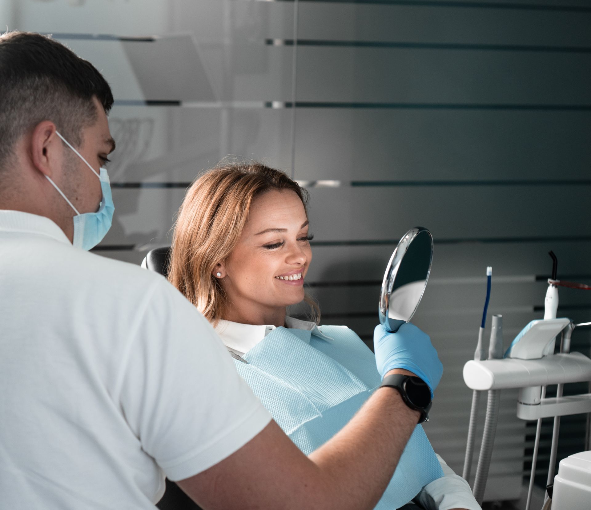 A woman is sitting in a dental chair looking at her teeth in a mirror.