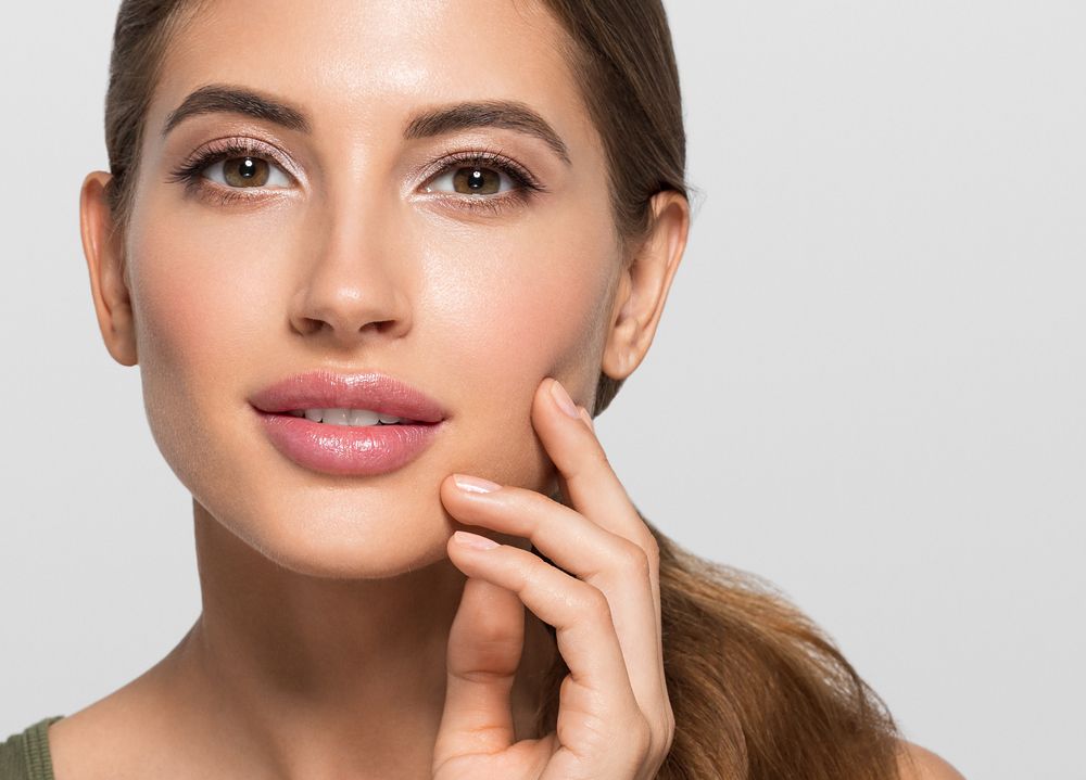 Close-up of a smiling woman with glossy lips and hand resting on her cheek against a light background