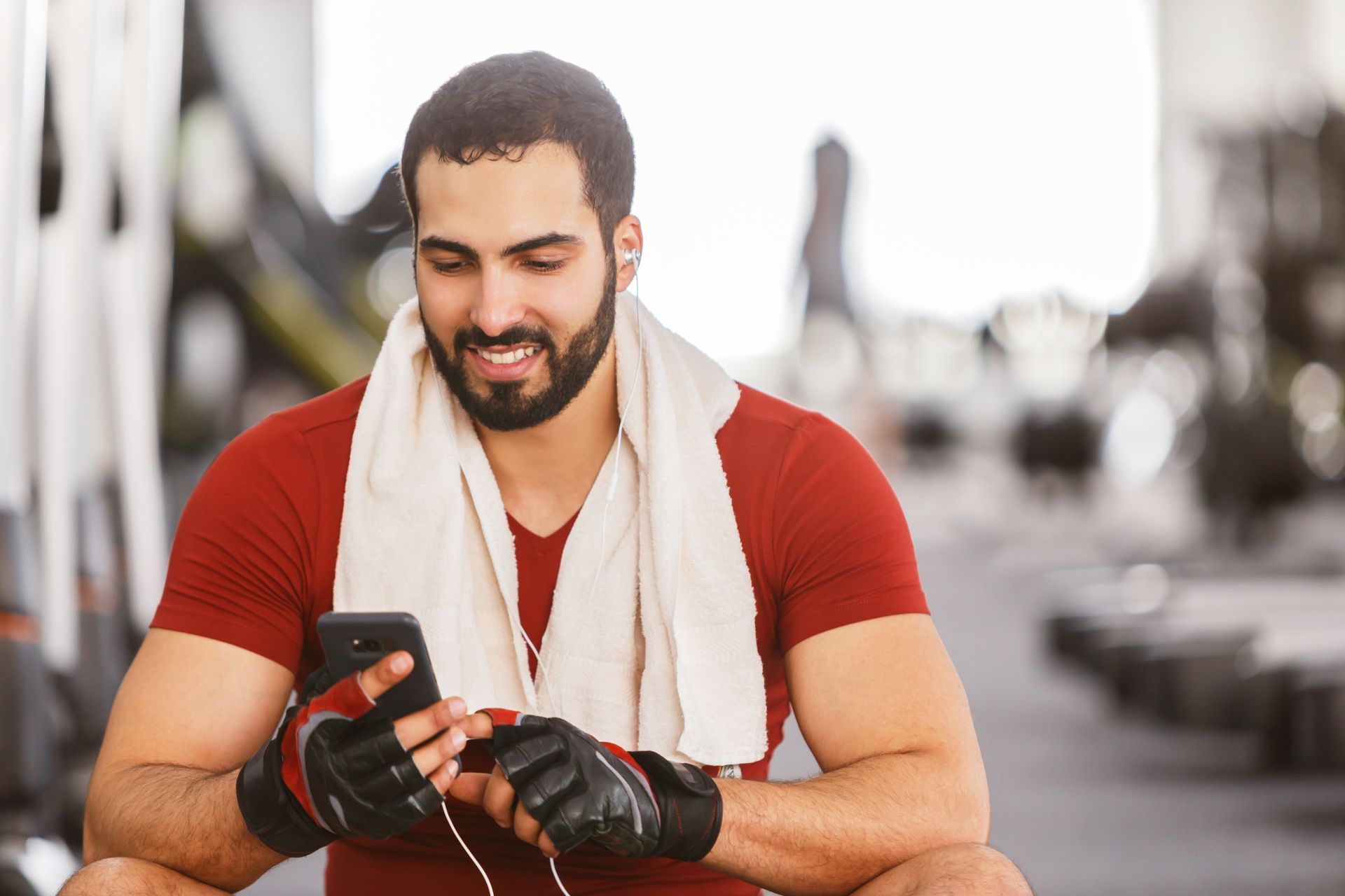 Man in red shirt at gym smiling, using a phone with a towel around his neck.