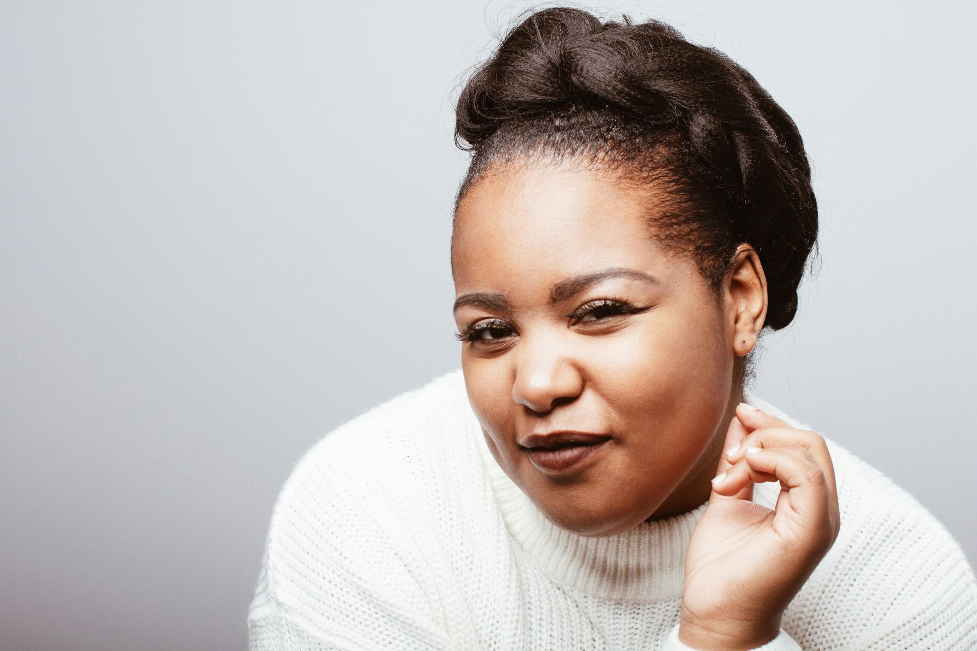 Woman with dark hair in a white sweater, looking and gesturing towards the viewer, with a gray background.
