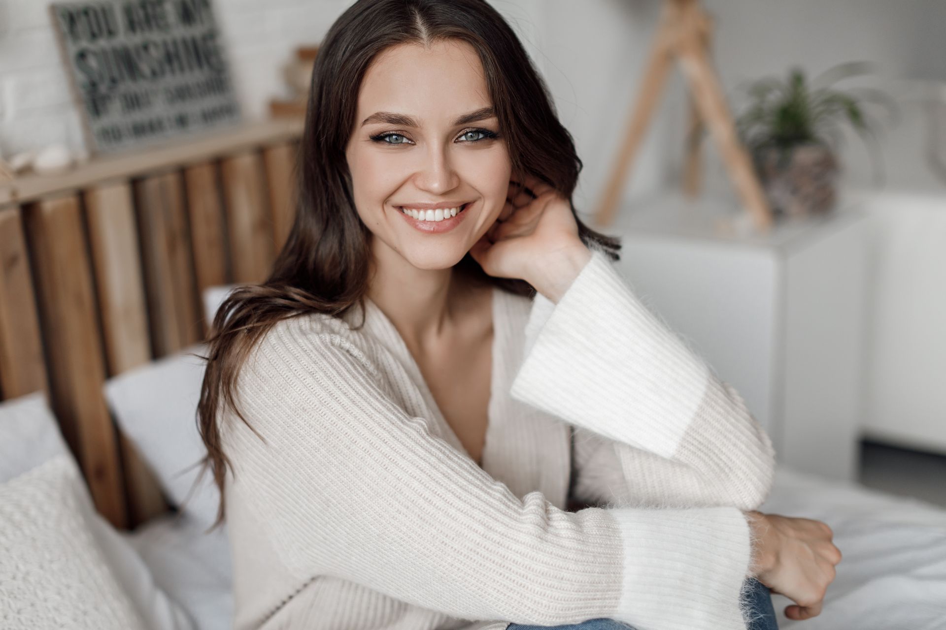 A woman is sitting on a bed wearing a white sweater and smiling.