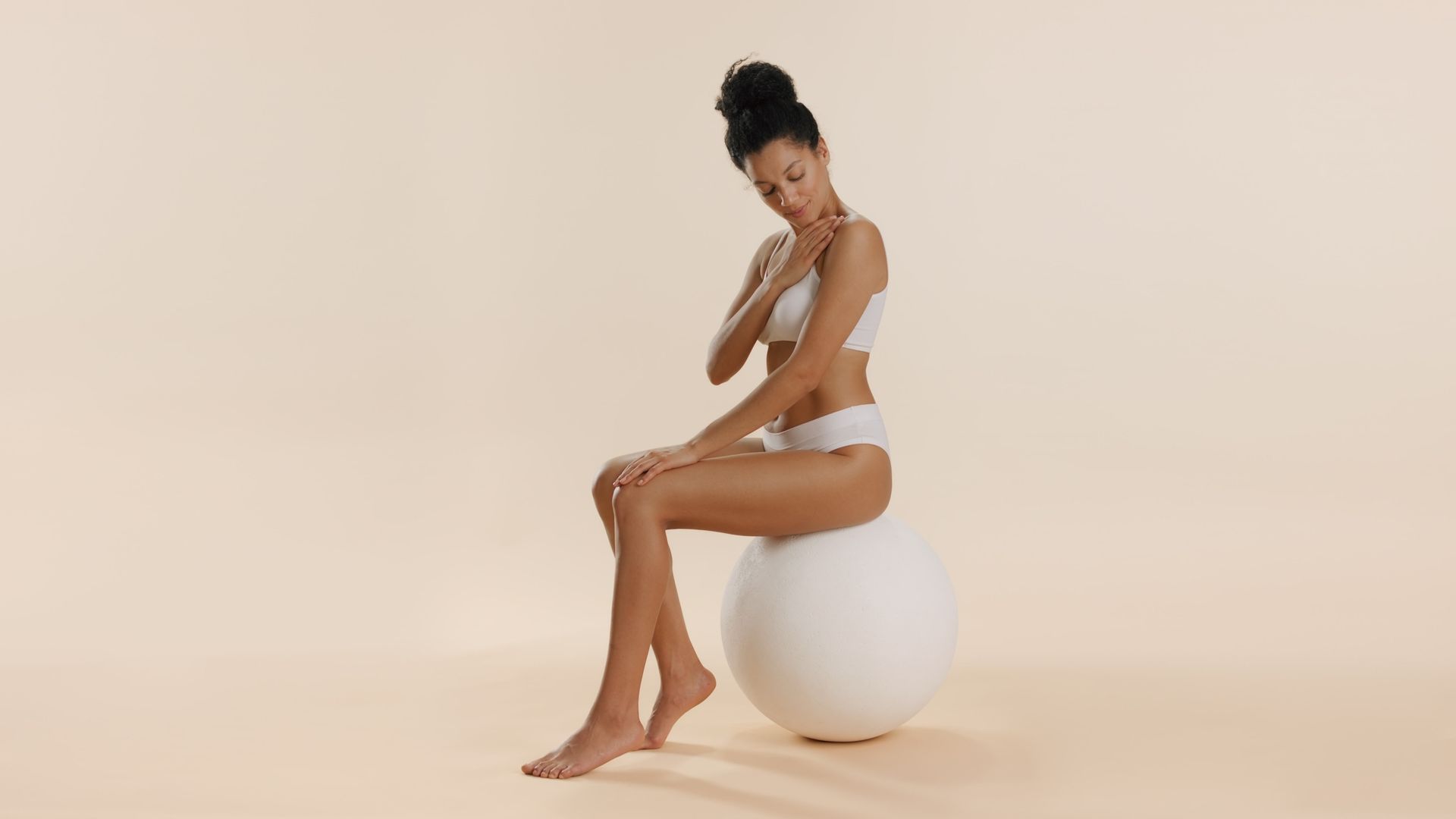 A person with hair in a bun wears matching white activewear while sitting on a large white exercise ball against a beige background.