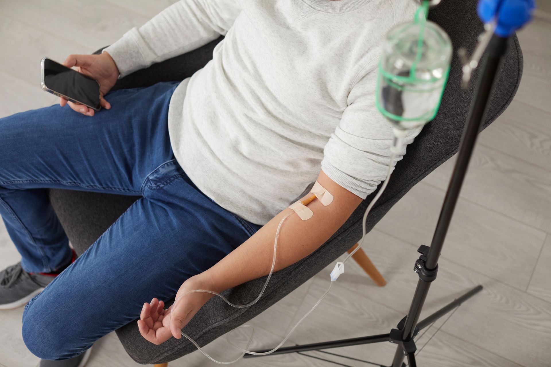 Person seated, receiving intravenous fluids. IV bag hanging from a stand, in a room.