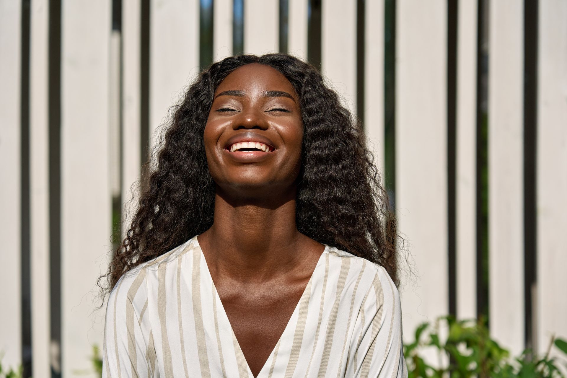 A woman is smiling with her eyes closed in front of a white fence.