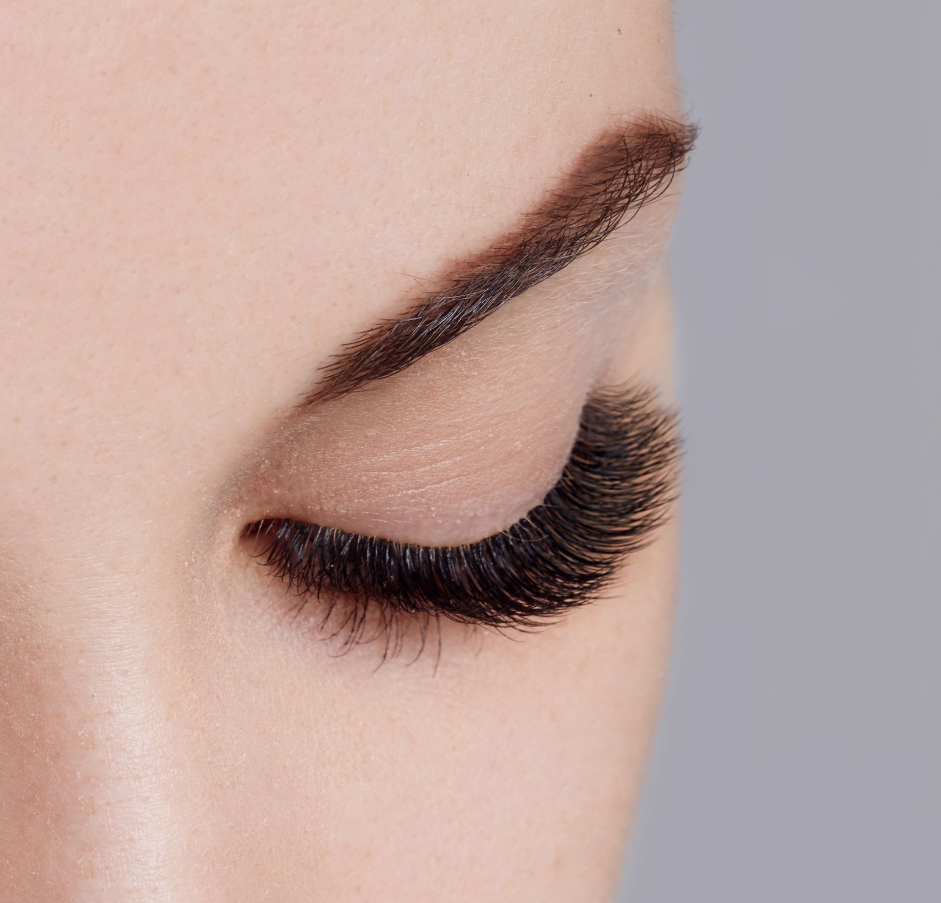 A close-up of a closed eye with long, dark, full eyelash extensions and a neatly groomed dark eyebrow.