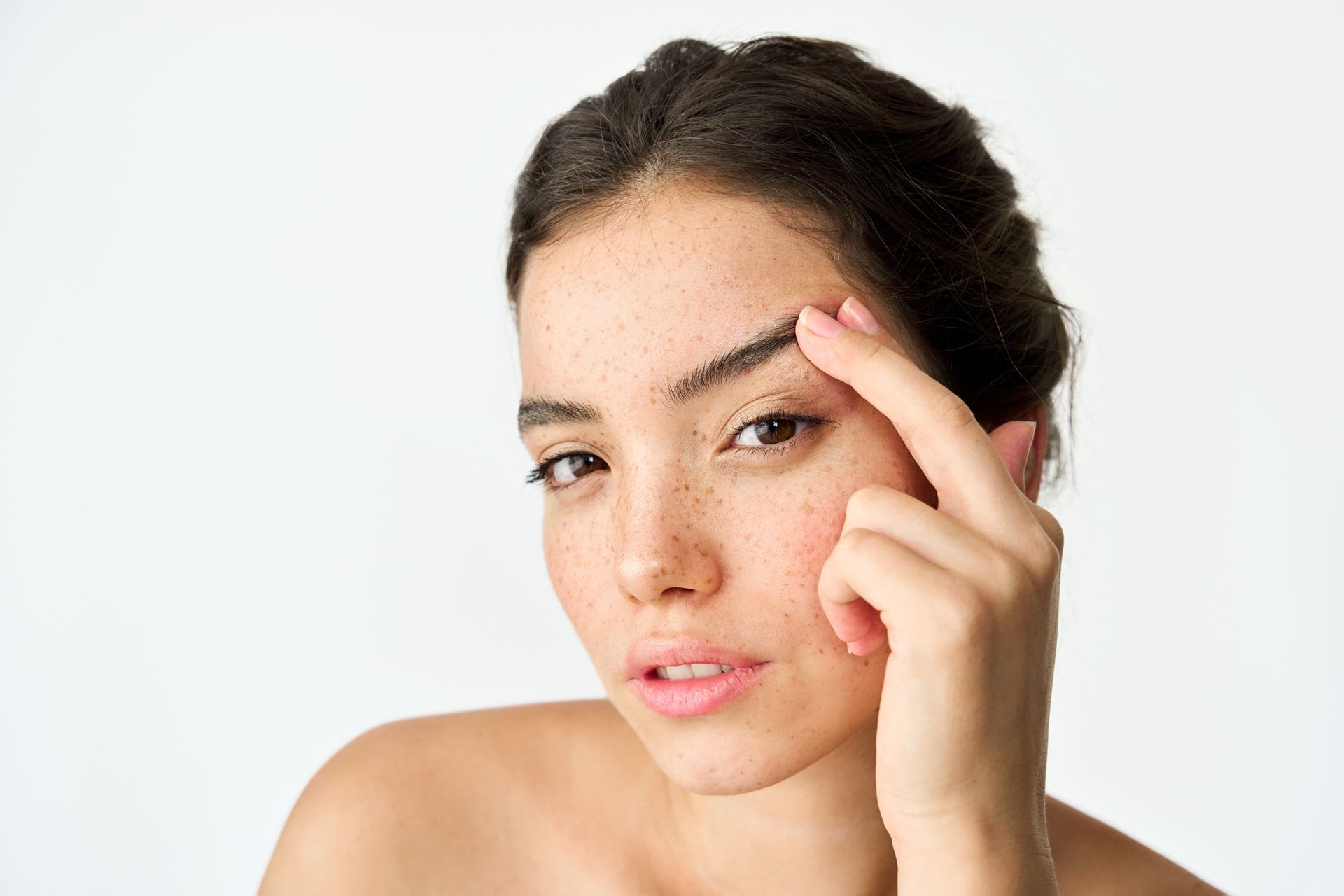 Woman touching eyebrow, looking at the viewer. Freckled skin, light background.
