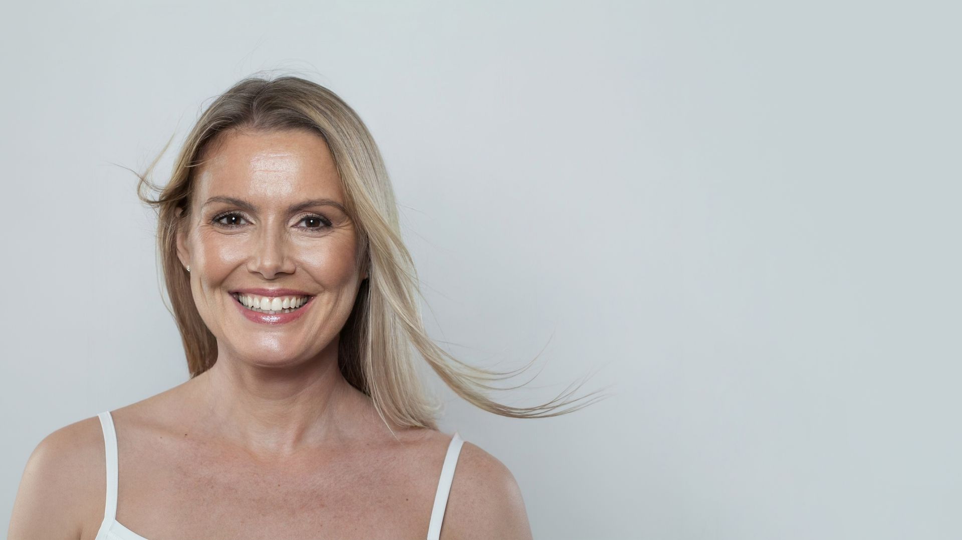 Woman with blonde hair smiling, wearing a white top, against a gray background.