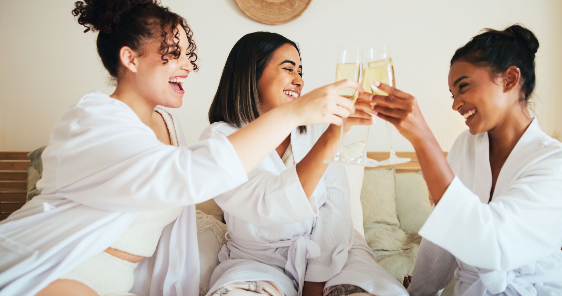 Three people in white robes toast with champagne glasses in a bedroom.