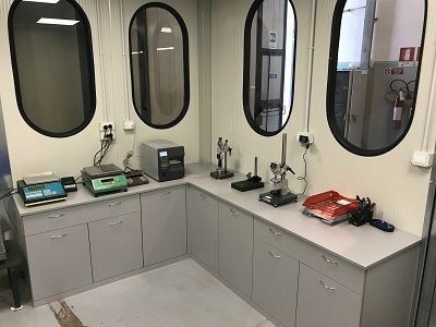 Sala di collaudo per apparecchiature elettroniche con strumenti a parete, contenitori e un piano di lavoro lungo alte finestre ovali.