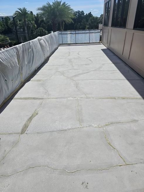 Concrete patio with cracks, enclosed by white barrier and building wall; trees and blue sky visible.