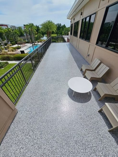 Outdoor patio with gray speckled flooring, metal railing, and white table, near a building with windows and chairs.