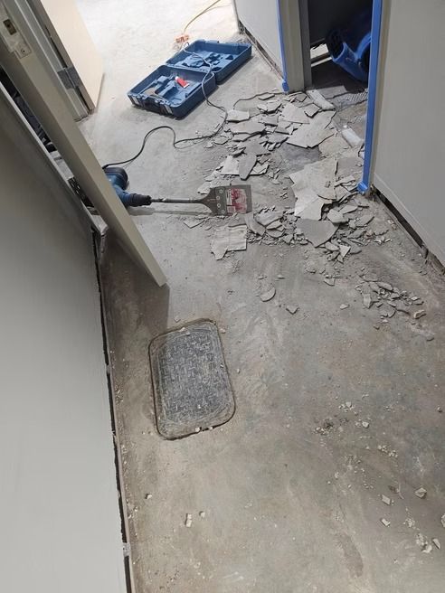 Demolition in hallway. Debris and jackhammer on concrete floor. Open doorway, tool box visible.
