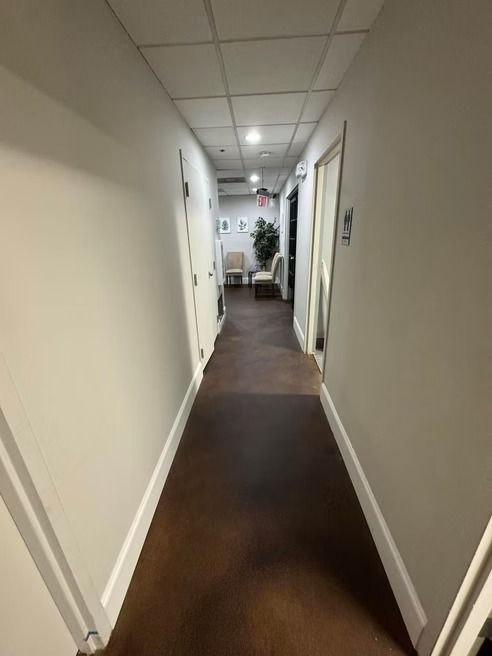 Narrow, brightly lit hallway with doors and a sitting area visible at the end. Dark floor, white walls and ceiling.