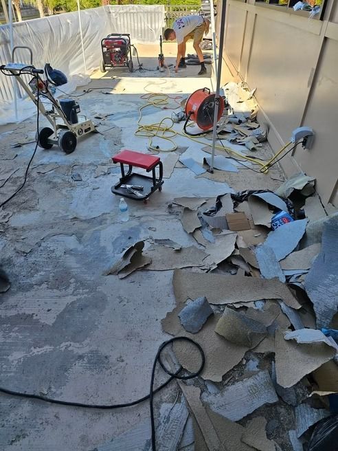 Workers removing flooring in a white-walled outdoor area, debris scattered around a bench, tools, and a fan present.
