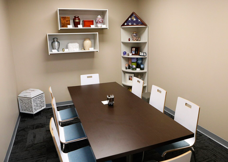 A small meeting room with a dark brown table, white chairs, and shelves displaying urns and a folded flag.