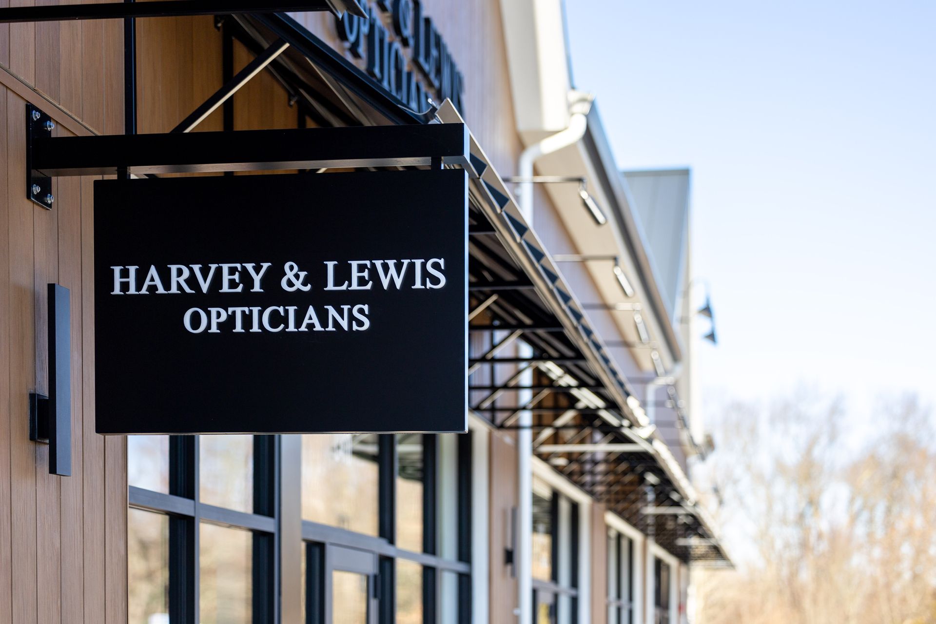 Sign for Harvey & Lewis Opticians, black on black, hanging outside a building with windows.