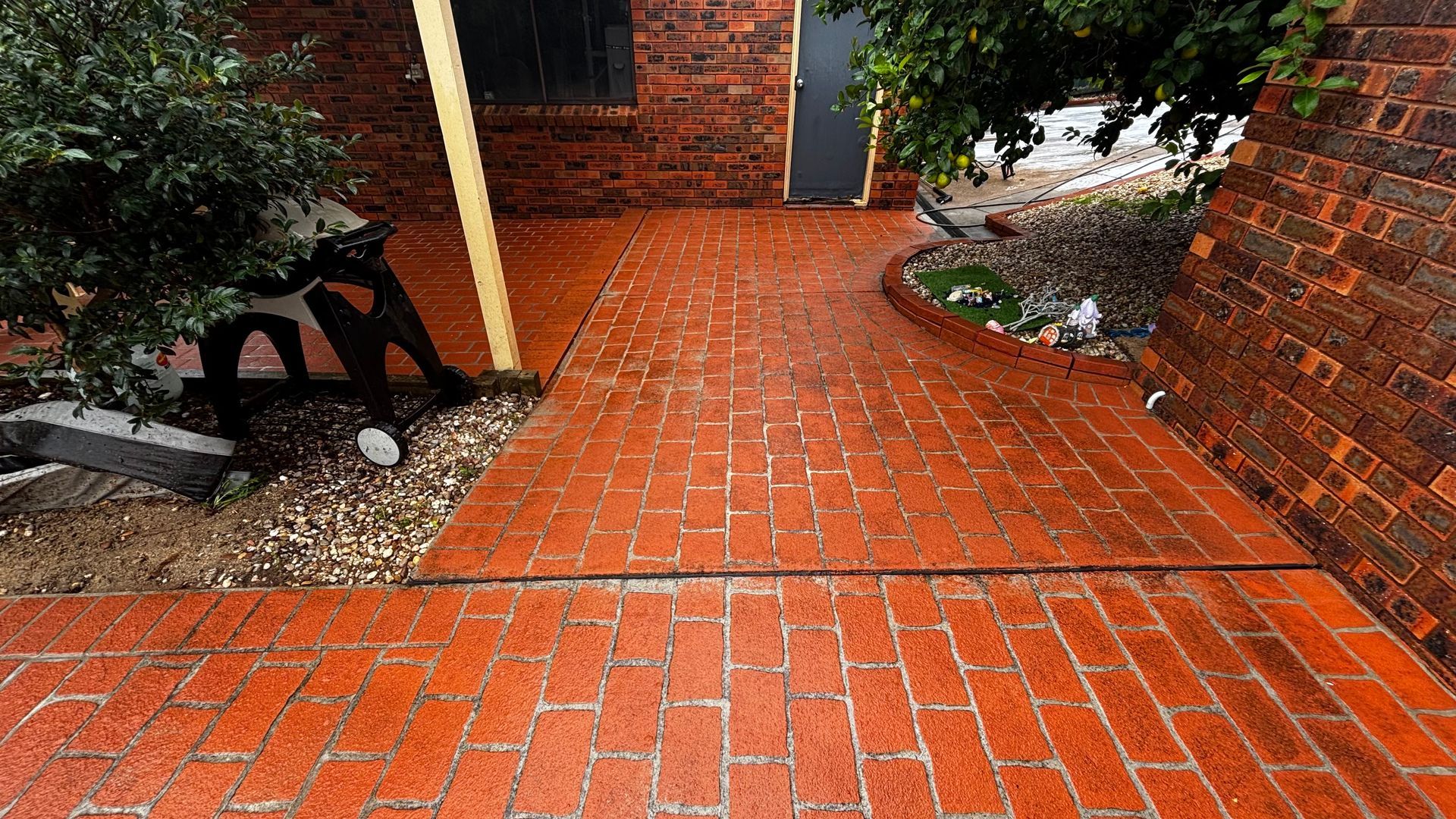 Brick walkway between brick walls, with a black bench on the left and garden plants nearby.