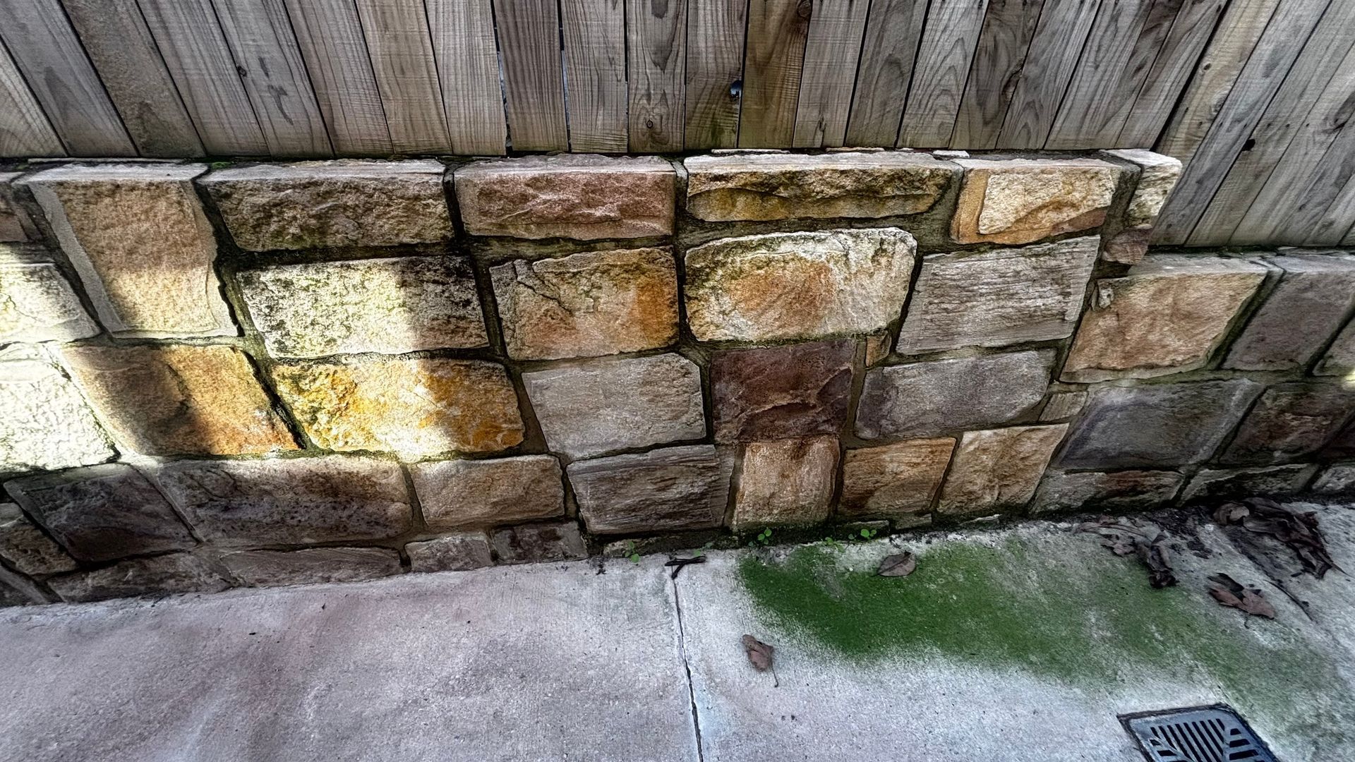 Stone retaining wall beside a wet concrete path with green moss and a drain grate