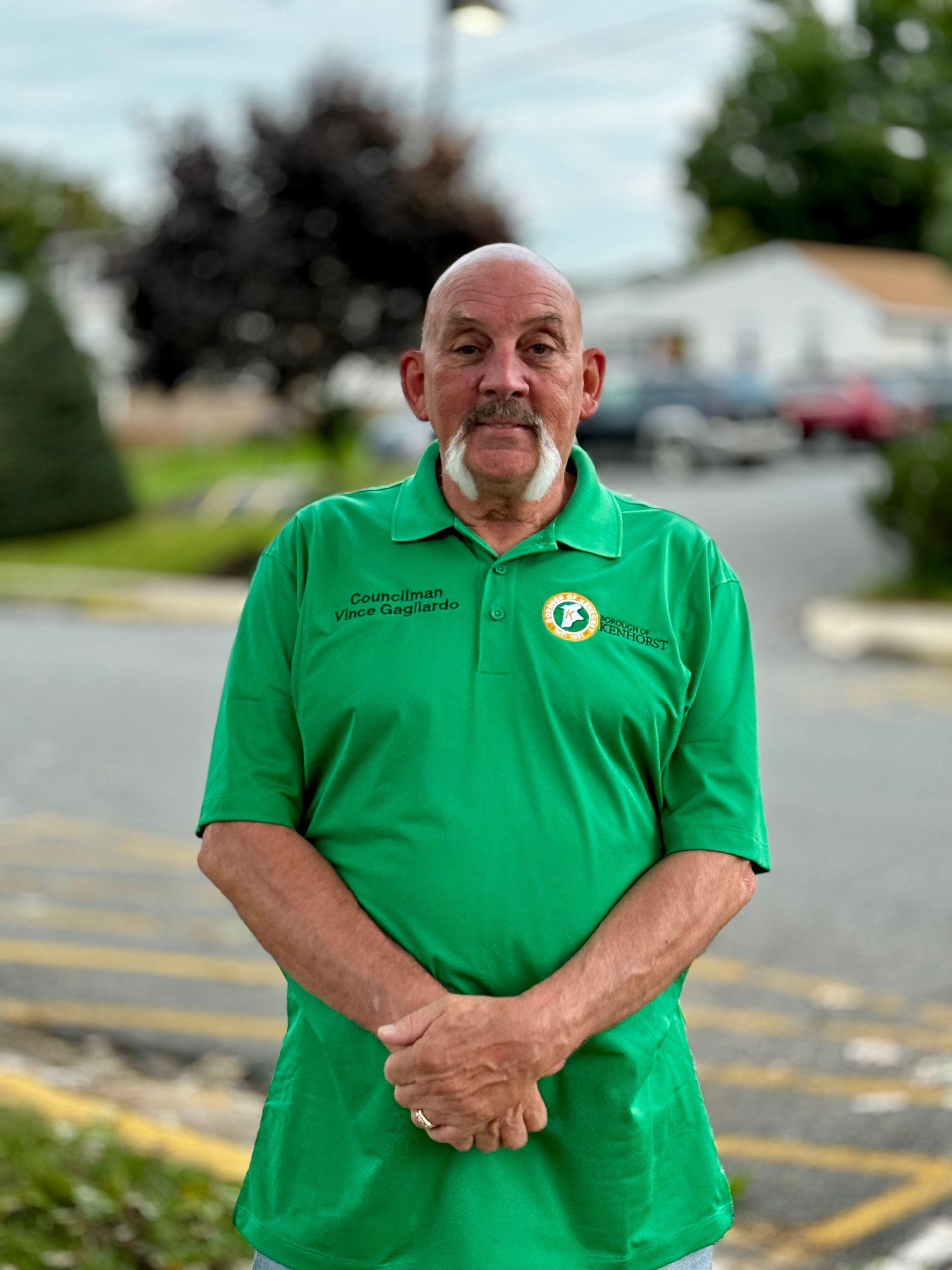 A man in a green shirt is standing in a parking lot with his hands folded.