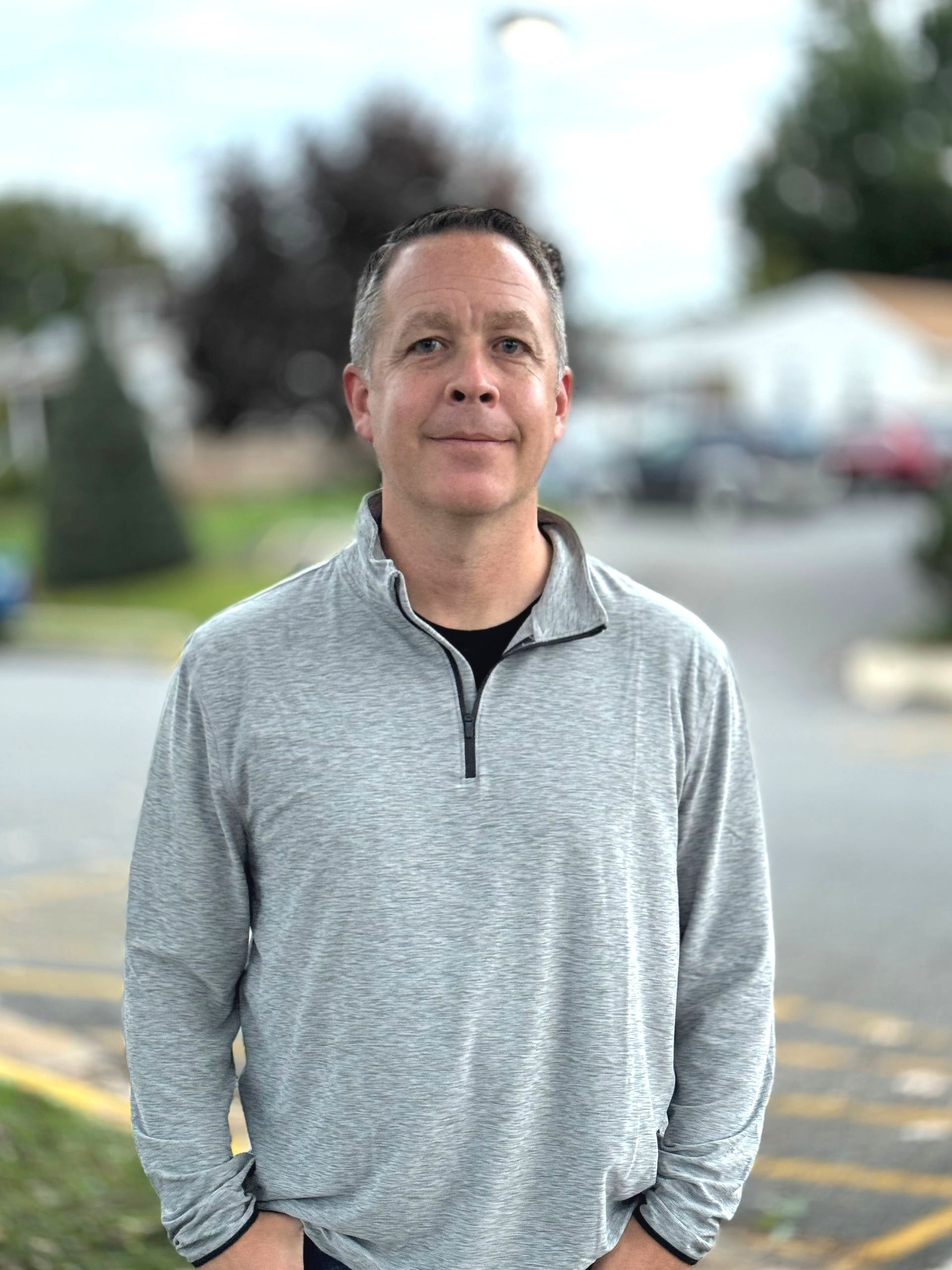 A man in a grey sweater is standing in a parking lot with his hands in his pockets.