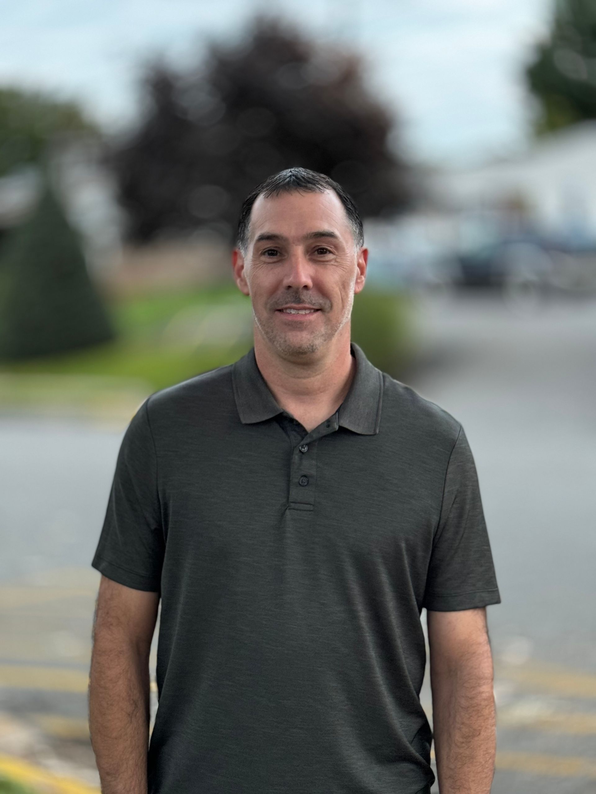A man in a grey polo shirt is standing in a parking lot.