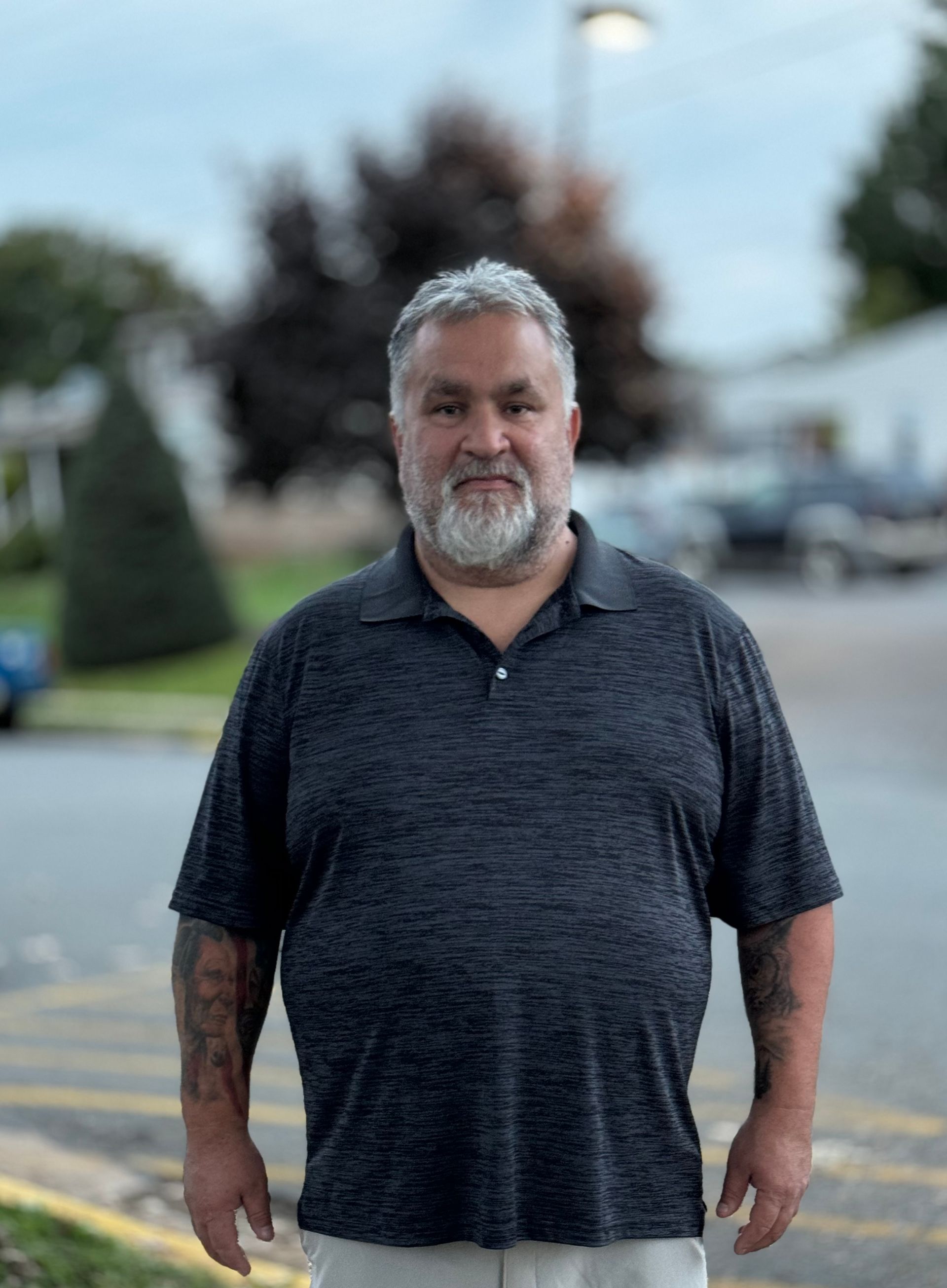 A man with a beard and tattoos is standing in a parking lot.
