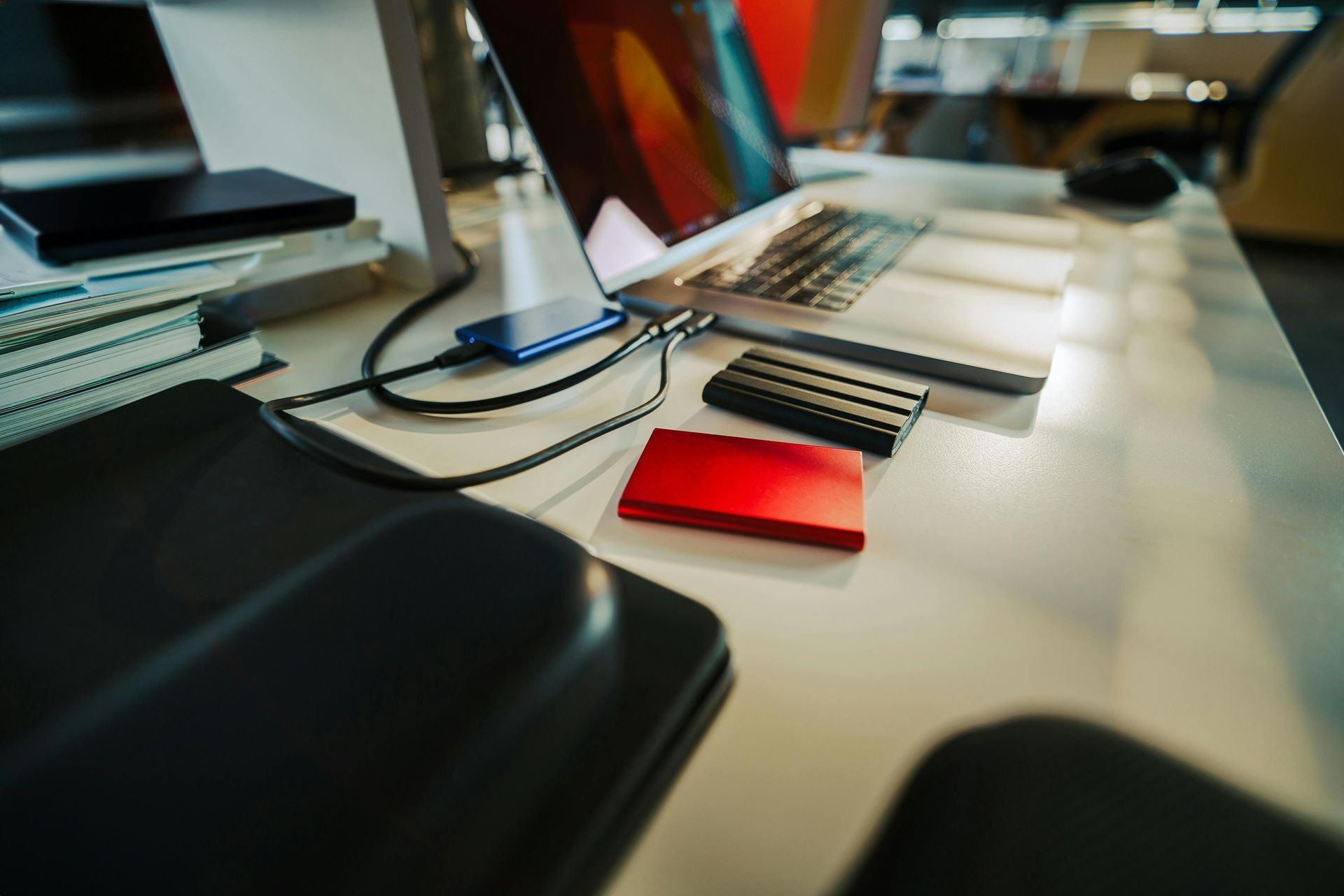 A laptop computer is sitting on a desk next to a red hard drive.