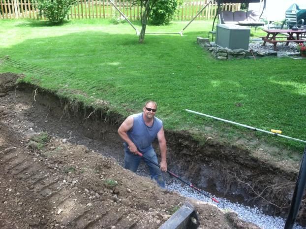 Man digging in a trench in a yard, filled with gravel, next to a grassy lawn.