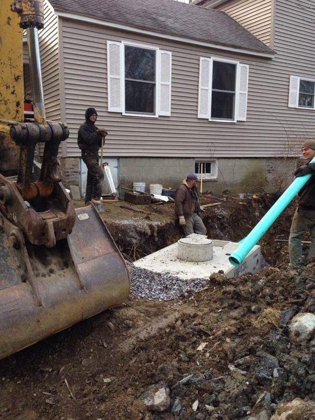 Construction workers installing a septic system. A backhoe is in foreground; men are digging and connecting pipes near a house.