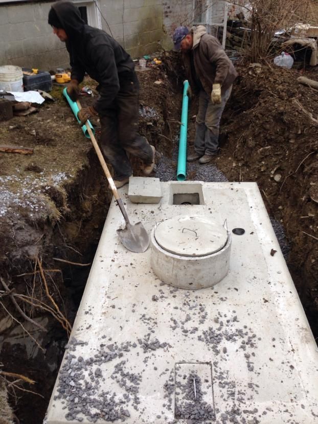 Two workers installing a concrete septic tank in a trench. One shovels while another connects a pipe.