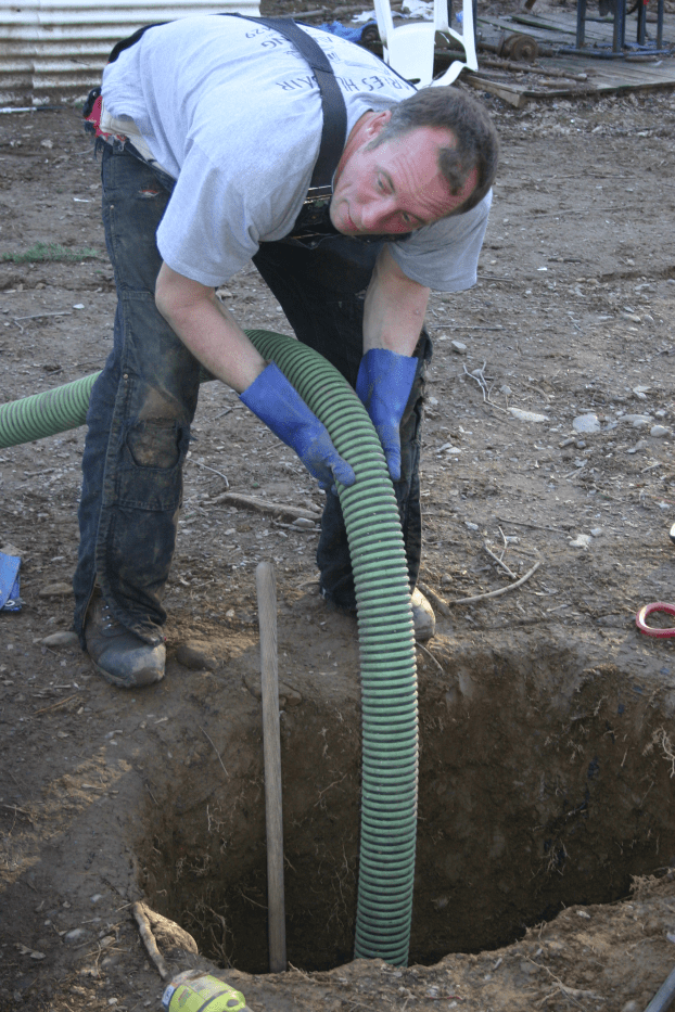 Man in overalls, bending over a hole, holding a green hose, blue gloves.